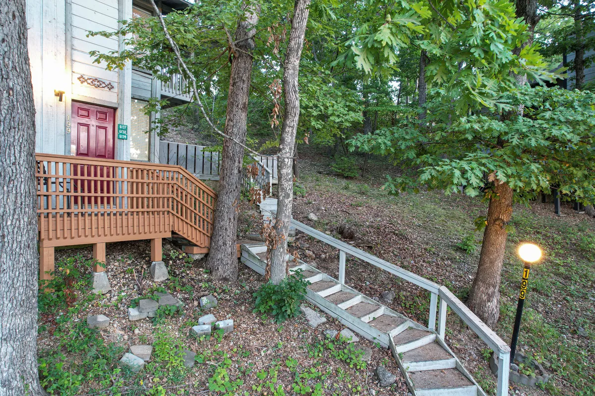 An inviting exterior featuring wooden stairs, an entry door, and tree-filled surroundings, with a pathway leading through the landscape. The natural light suggests a serene atmosphere ideal for nature walks and sunset views.