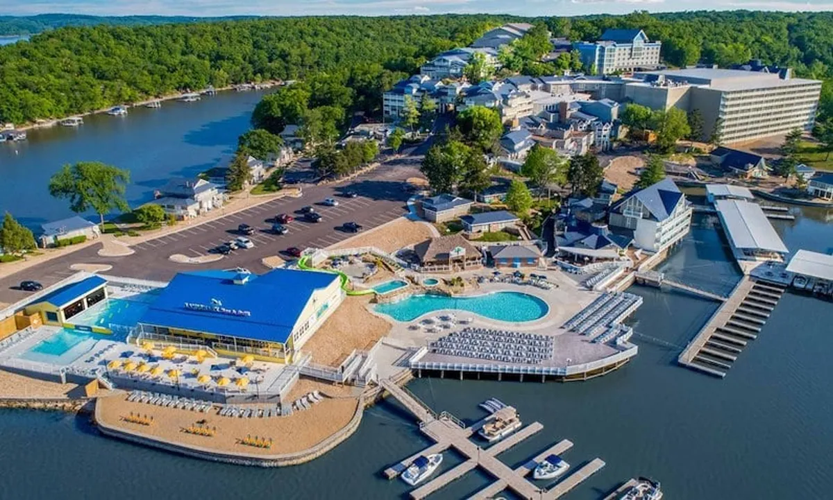 The image showcases a vibrant resort setting at sunset, featuring a sparkling swimming pool bordered by a sandy beach area, boats docked nearby on the lake, and the inviting hotel complex in the background. Guests are relaxing poolside, enjoying the water, and soaking in the warm, leisurely atmosphere with easy access to both aquatic adventures and laid-back lounging.