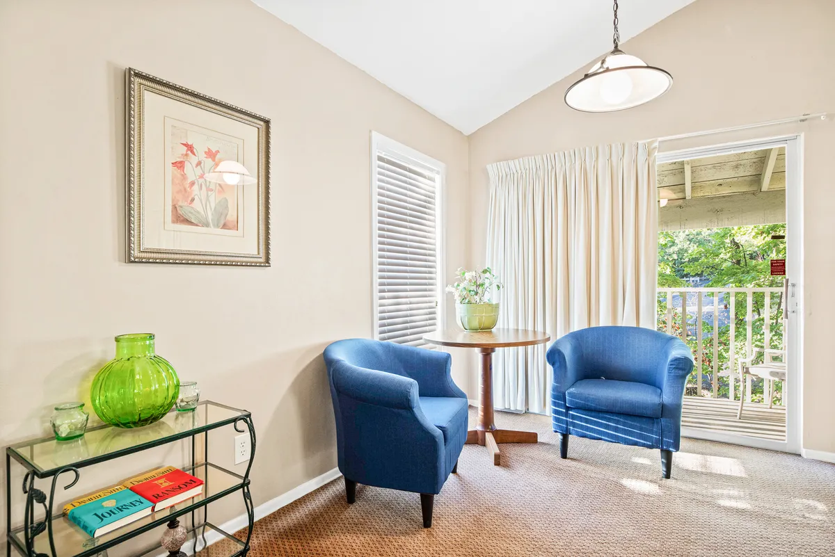 A cozy indoor reading nook with two blue chairs positioned near a table topped with books and a decorative green vase. Sunlight pours in, creating a perfect spot for conversation, meditation, or enjoying a good book. The inviting setup encourages restful moments and personal reflection.