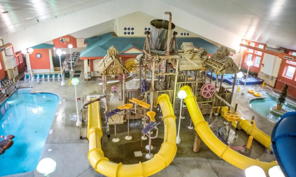 An energetic indoor scene showing a vibrant pool area with twisting water slides, play structures, and families enjoying the water. The bright, joyful atmosphere captures the essence of resort-style amenities and fun for children and adults alike.