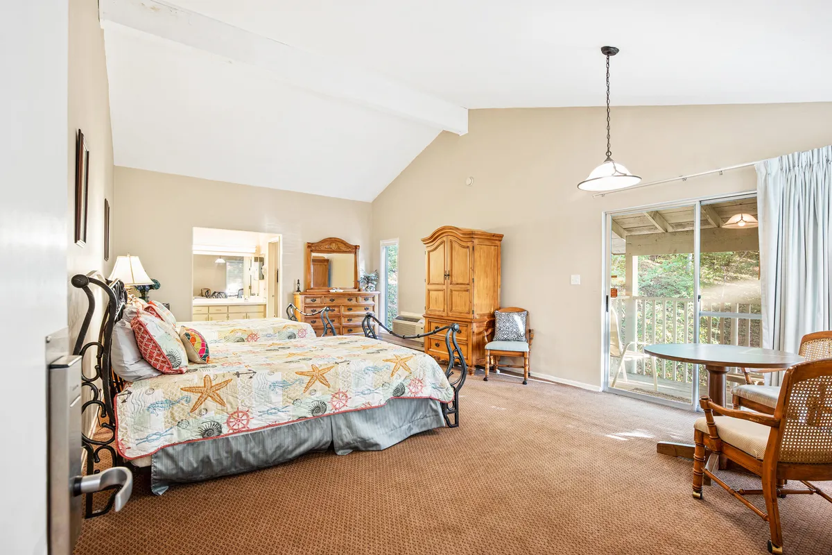A comfortable, inviting bedroom with two beds dressed in decorative quilts, a wooden dresser, and a door leading to a private balcony. The space is bathed in natural light, with direct access to an outdoor area ideal for morning coffee or evening reflection. The overall ambiance is restful and welcoming.