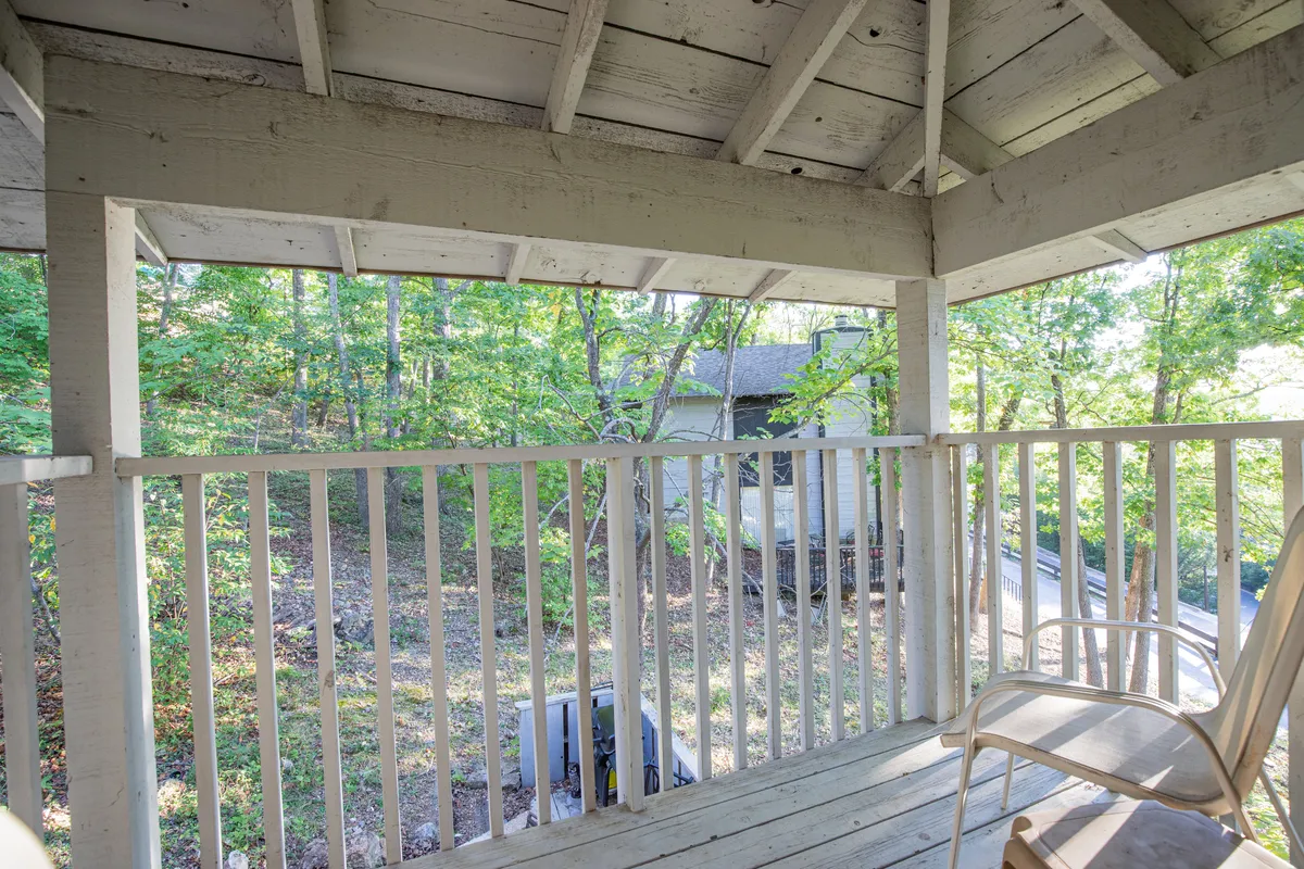 A view from a wooden balcony or deck surrounded by lush trees, with a wooden railing and an adjacent structure visible. The scene suggests relaxation, quiet moments, and a seamless connection to the outdoors.