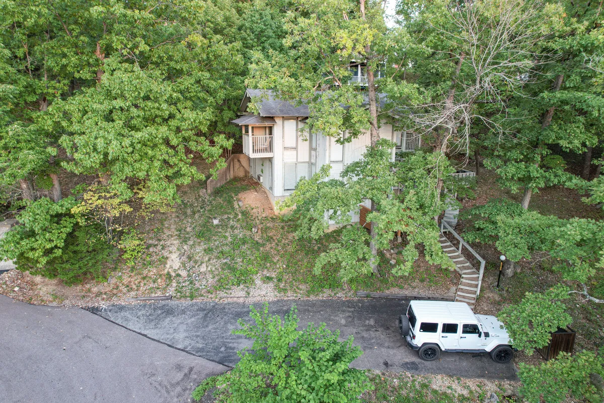 A peaceful exterior scene showing the house nestled among mature trees, with a winding pathway leading up to it and a white jeep parked nearby. The setting emphasizes seclusion, serenity, and immersion in nature.