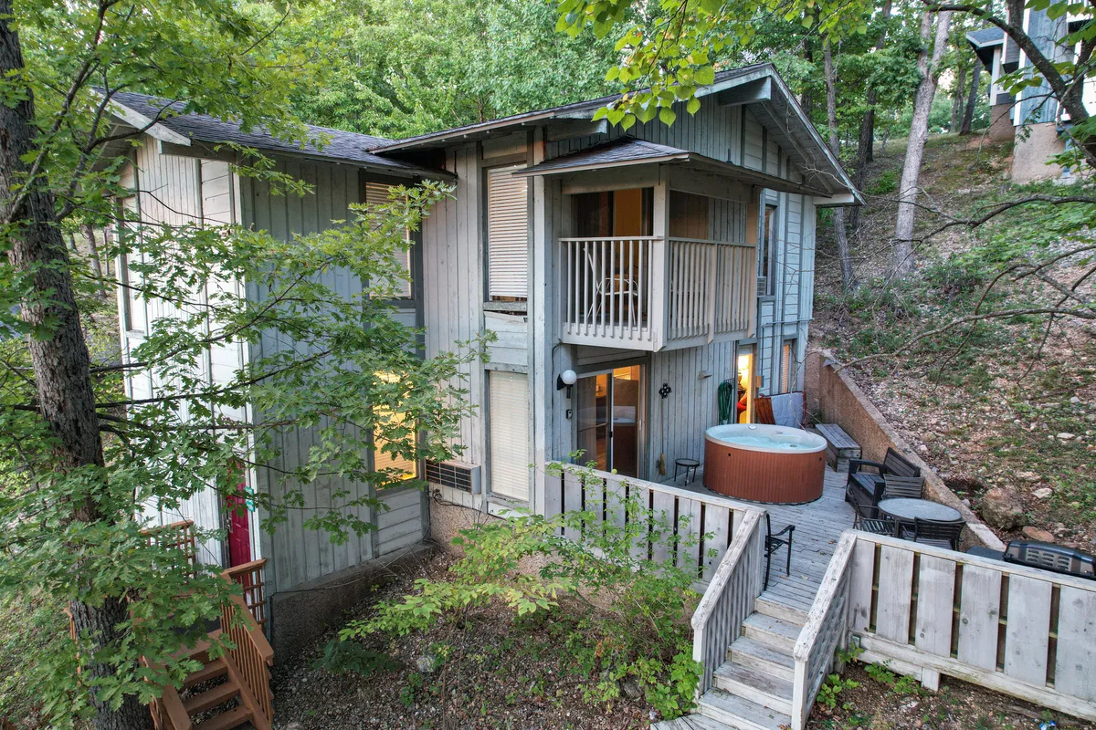 An inviting exterior shot featuring wooden steps leading up to a spacious balcony, a visible hot tub, large windows, and a backdrop of dense trees. The scene hints at activities such as relaxing in the hot tub, enjoying a meal outdoors, or stargazing from the balcony.