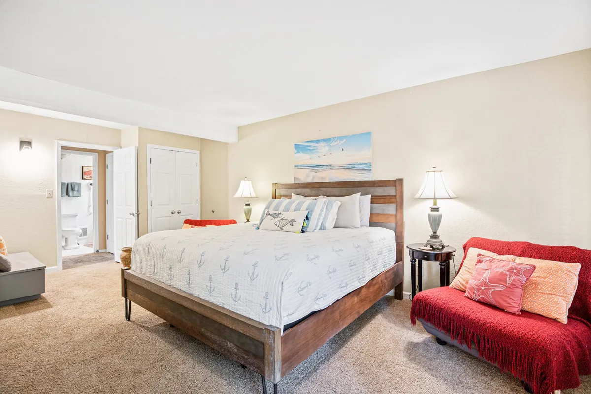 A tranquil bedroom featuring a king-sized bed dressed in nautical-themed bedding, highlighted by soft natural light streaming in. Above the bed is calming beach artwork, while a vibrant red accent chair offers a cozy nook for reading or quiet conversation. The scene radiates warmth and comfort, inviting relaxation at any time of day.