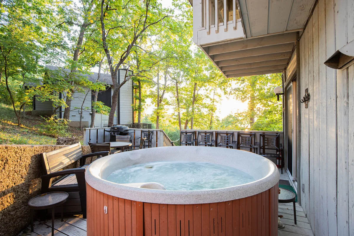 A tranquil outdoor space featuring a bubbling hot tub surrounded by lush trees, a spacious deck with comfortable seating, and a view designed for relaxation and gathering. The area is bathed in soft natural light, inviting guests to unwind or socialize in the serene woodland setting.