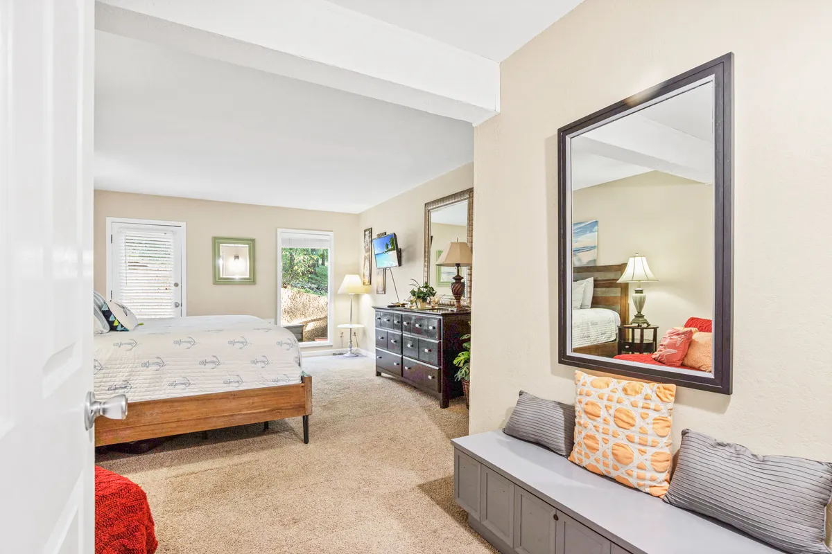A cozy bedroom scene with a comfortable bed dressed in decorative pillows, a large mirror, dresser, and natural light flooding through the window. The space embodies relaxation and a sense of being cared for, encouraging moments of rest and tranquility.